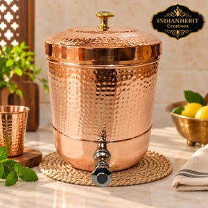 Könnte beinhalten: Ein zylindrischer Wasserspender aus Kupfer mit gehämmertem Muster, einem abnehmbaren Deckel mit goldenem Knauf und einem Metallhahn an der Basis. Die Oberfläche reflektiert das Licht und das Logo INDIANHERIT Creations ist oben rechts zu sehen.