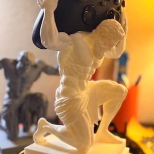 May include: A white 3D-printed statue of a kneeling figure with a muscular build, positioned on a square base. The figure holds a black wireless gaming controller above its head with both arms. A second, similar grey statue is visible in the background.