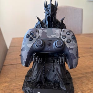 Puede incluir: Una figura negra de un personaje encapuchado y blindado con una corona de pinchos que sostiene un mando de videojuegos con estampado de camuflaje gris y negro. La base tiene una placa negra con el texto blanco PS5 impreso en el frente.