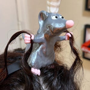May include: A gray rat figurine wearing a white chef hat, positioned on a dark headband. The rat has pink paws and a pink nose, and its front paws are holding two strands of dark brown hair. The figure has a smooth, layered texture.