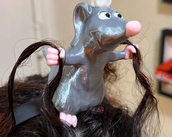 May include: A gray rat figurine wearing a white chef hat, positioned on a dark headband. The rat has pink paws and a pink nose, and its front paws are holding two strands of dark brown hair. The figure has a smooth, layered texture.
