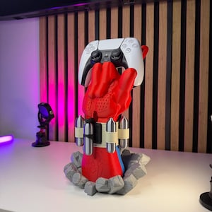 May include: A red and blue superhero-themed controller stand shaped like a gloved hand emerging from a gray rocky base. The hand features a beige wrist strap with four silver canisters and holds a white and black wireless gaming controller.