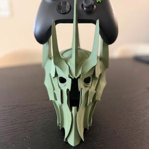 May include: A matte light green 3D printed stand shaped like a pointed, jagged helmet with a central spike, supporting a black wireless video game controller with colored buttons on the top, featuring a symmetrical, angular, and sharp design.