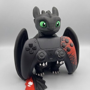 Puede incluir: Una figura de dragón negro con ojos verde brillante que sostiene un mando de juego negro y rojo. El mando presenta una empuñadura texturizada y un patrón gráfico rojo en el lado derecho. El dragón tiene pequeñas garras blancas y una aleta roja.