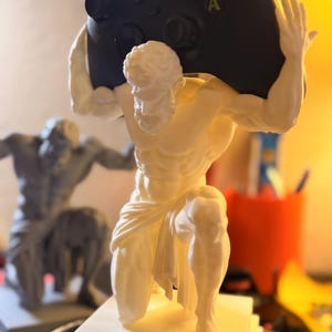 Puede incluir: Una escultura blanca de una figura masculina arrodillada que sostiene un mando de videojuego negro sobre su cabeza. El mando tiene botones Y, X, B y A visibles. La figura está colocada sobre una base cuadrada, con una escultura gris similar visible al fondo.