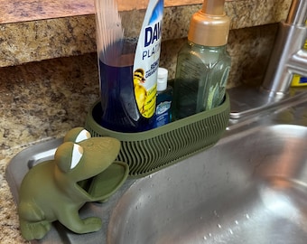 Può includere: Un porta spugna a forma di rana color verde oliva con occhi bianchi e bocca aperta, posizionato accanto a un contenitore ovale a coste che ospita una bottiglia di detersivo blu, un piccolo igienizzante mani e un dispenser di sapone verde.