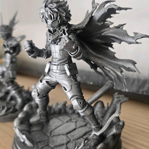May include: A metallic silver-colored figurine of a character in a dynamic pose with a flowing cape, standing on a textured circular base. The figure has spiky hair, a fitted suit with utility pouches, and is positioned with one fist raised forward.