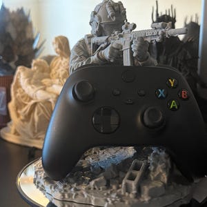 May include: A black video game controller rests on a gray 3D-printed stand featuring a soldier in tactical gear aiming a rifle. The soldier stands atop a base of rubble and debris. The controller features standard buttons labeled Y, X, B, and A.