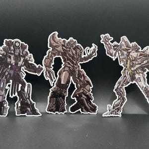 Könnte beinhalten: Drei gestanzte Aufkleber mit detaillierten Illustrationen von Roboterfiguren. Die linke Figur ist dunkelgrau mit mechanischen Details, die mittlere Figur ist ein braun getönter gepanzerter Roboter und die rechte Figur ist ein hellgrauer Roboter mit Flügeln.