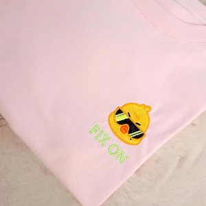 Op de afbeelding: Een lichtroze t-shirt met een geborduurd geel kuikentje dat een zwarte zonnebril draagt. Onder het kuikentje is de tekst FIX ON geborduurd met lichtgroene draad. Het kledingstuk heeft een ronde hals en korte mouwen, in een gevouwen lay-out.