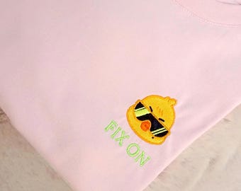 Puede incluir: Una camiseta rosa claro con un bordado de un pollito amarillo que lleva gafas de sol negras. Debajo del pollito, el texto FIX ON está bordado en hilo verde claro. La prenda tiene cuello redondo y mangas cortas, presentada doblada.