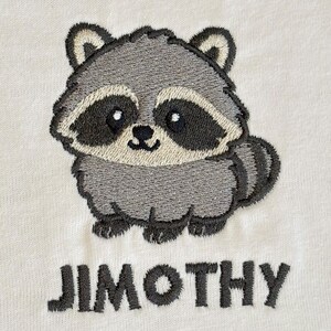 Puede incluir: Una tela de color blanco roto con un dise&ntilde;o bordado de un mapache de dibujos animados con pelaje gris, un antifaz negro y una cola a rayas. Debajo del mapache, el nombre JIMOTHY est&aacute; bordado en letras may&uacute;sculas de color gris oscuro.