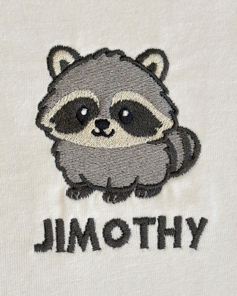 Puede incluir: Una tela de color blanco roto con un dise&ntilde;o bordado de un mapache de dibujos animados con pelaje gris, un antifaz negro y una cola a rayas. Debajo del mapache, el nombre JIMOTHY est&aacute; bordado en letras may&uacute;sculas de color gris oscuro.