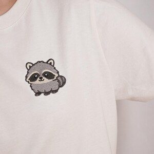 Op de afbeelding: Een wit T-shirt met korte mouwen en een kleine, geborduurde afbeelding van een grijze en zwarte wasbeer op de linkerborst. Het wasbeerontwerp heeft grote ogen, een gestreepte staart en een lichtgrijs gezicht met donkere oogvlekken.