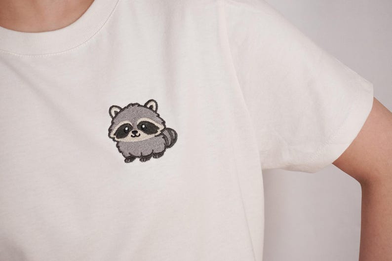 Op de afbeelding: Een wit T-shirt met korte mouwen en een kleine, geborduurde afbeelding van een grijze en zwarte wasbeer op de linkerborst. Het wasbeerontwerp heeft grote ogen, een gestreepte staart en een lichtgrijs gezicht met donkere oogvlekken.