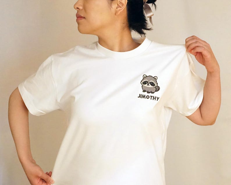 Puede incluir: Una camiseta blanca de manga corta con un peque&ntilde;o gr&aacute;fico de un mapache de dibujos animados en la parte izquierda del pecho. Debajo de la ilustraci&oacute;n del mapache, el nombre JIMOTHY est&aacute; impreso en letras may&uacute;sculas de color marr&oacute;n oscuro.