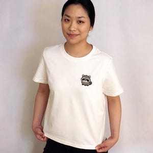 Op de afbeelding: Een cr&egrave;mekleurig T-shirt met korte mouwen en een kleine, geborduurde afbeelding van een schattige, ronde wasbeer op de linkerborst. Het shirt heeft een klassieke ronde hals en een rechte zoom voor een eenvoudige en strakke uitstraling.