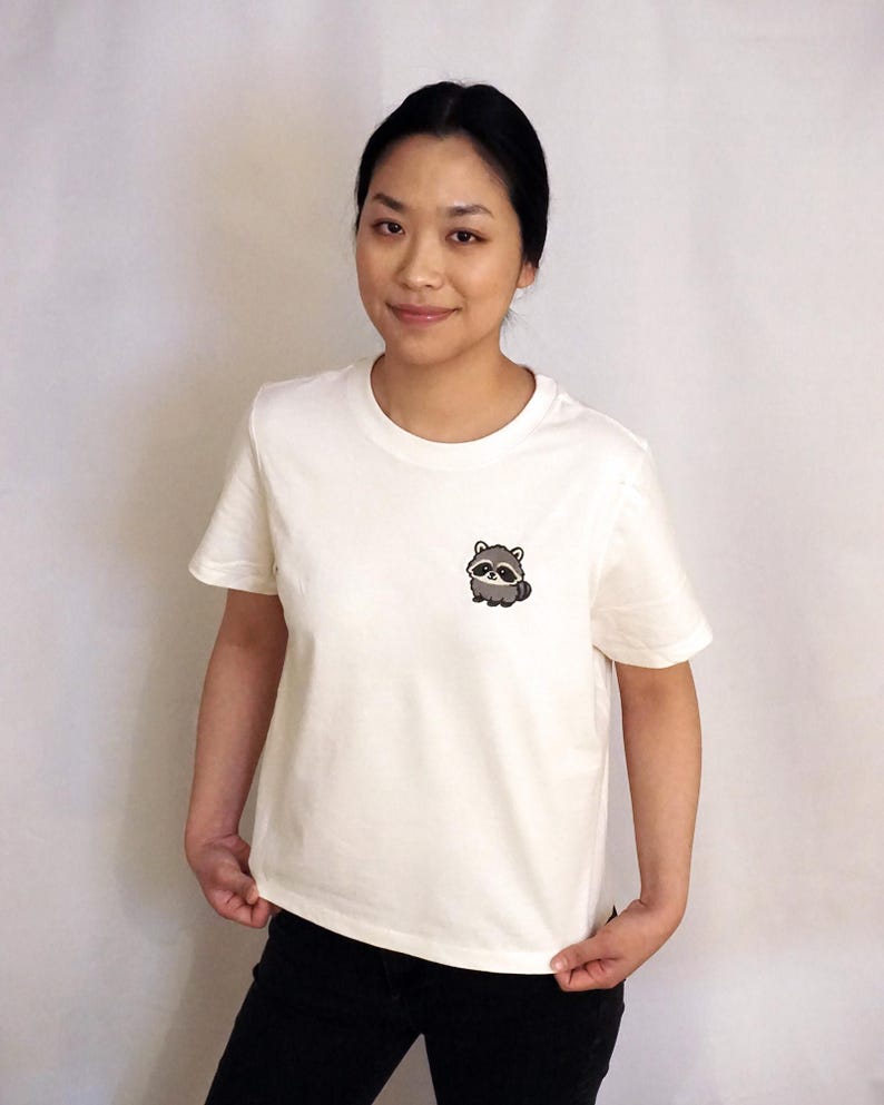 Op de afbeelding: Een cr&egrave;mekleurig T-shirt met korte mouwen en een kleine, geborduurde afbeelding van een schattige, ronde wasbeer op de linkerborst. Het shirt heeft een klassieke ronde hals en een rechte zoom voor een eenvoudige en strakke uitstraling.