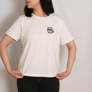 Op de afbeelding: Een off-white T-shirt met korte mouwen en een ronde hals, voorzien van een kleine, geborduurde afbeelding van een cartoon wasbeer op de linkerborst. Het shirt heeft een standaard pasvorm met een rechte zoom en een donkere broek.