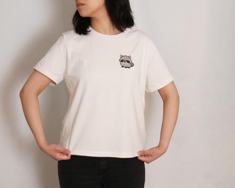 Op de afbeelding: Een off-white T-shirt met korte mouwen en een ronde hals, voorzien van een kleine, geborduurde afbeelding van een cartoon wasbeer op de linkerborst. Het shirt heeft een standaard pasvorm met een rechte zoom en een donkere broek.