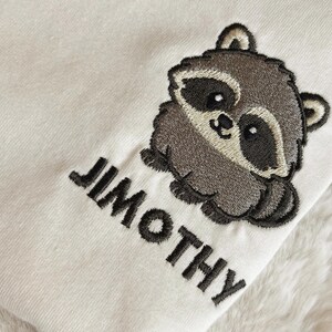 Puede incluir: Una tela de color crema con un dise&ntilde;o bordado de un peque&ntilde;o mapache redondo con pelaje gris oscuro y marr&oacute;n claro. Debajo del mapache, el nombre JIMOTHY est&aacute; bordado en letras may&uacute;sculas de color gris oscuro con una fuente sans-serif.