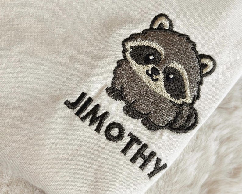 Puede incluir: Una tela de color crema con un dise&ntilde;o bordado de un peque&ntilde;o mapache redondo con pelaje gris oscuro y marr&oacute;n claro. Debajo del mapache, el nombre JIMOTHY est&aacute; bordado en letras may&uacute;sculas de color gris oscuro con una fuente sans-serif.