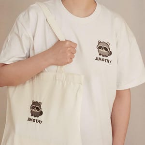 Puede incluir: Una camiseta blanca de manga corta y una bolsa de tela blanca a juego, ambas con un peque&ntilde;o gr&aacute;fico de un mapache de dibujos animados y el texto "JIMOTHY" impreso en marr&oacute;n oscuro. El mapache tiene pelaje gris y marr&oacute;n.
