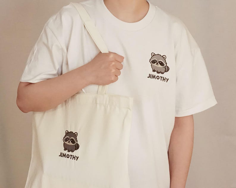 Puede incluir: Una camiseta blanca de manga corta y una bolsa de tela blanca a juego, ambas con un peque&ntilde;o gr&aacute;fico de un mapache de dibujos animados y el texto "JIMOTHY" impreso en marr&oacute;n oscuro. El mapache tiene pelaje gris y marr&oacute;n.