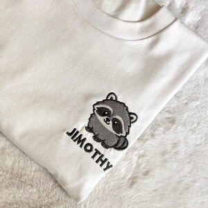 Puede incluir: Una camiseta de color crema con un dise&ntilde;o bordado en el pecho. El bordado muestra un peque&ntilde;o mapache gris de estilo caricatura con manchas negras alrededor de los ojos y el nombre JIMOTHY bordado en letras may&uacute;sculas negras debajo.