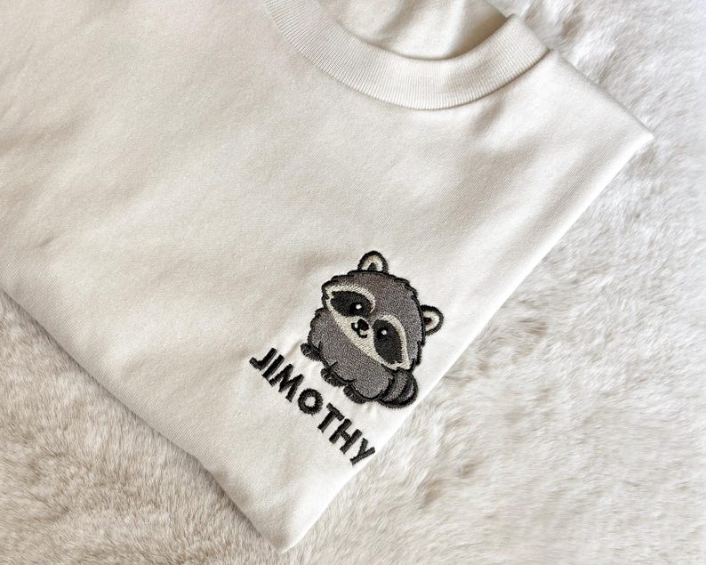 Puede incluir: Una camiseta de color crema con un dise&ntilde;o bordado en el pecho. El bordado muestra un peque&ntilde;o mapache gris de estilo caricatura con manchas negras alrededor de los ojos y el nombre JIMOTHY bordado en letras may&uacute;sculas negras debajo.