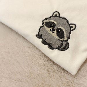 Op de afbeelding: Een wit kledingstuk van stof met een kleine, geborduurde wasbeer. De wasbeer is afgebeeld in grijze, zwarte en witte tinten met een ronde, cartoonachtige uitstraling, grote donkere ogen en een gestreepte staart op de stof.