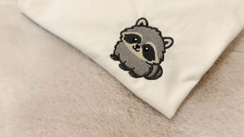 Op de afbeelding: Een wit kledingstuk van stof met een kleine, geborduurde wasbeer. De wasbeer is afgebeeld in grijze, zwarte en witte tinten met een ronde, cartoonachtige uitstraling, grote donkere ogen en een gestreepte staart op de stof.