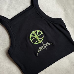 Op de afbeelding: Een zwart geribbelde tanktop met een cirkelvormige limoengroene geborduurde afbeelding op de borst. Onder de cirkel bevindt zich een extra limoengroen geborduurd ontwerp met gestileerde, scherpe en hoekige letters.