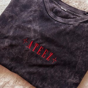 Op de afbeelding: Een zwart acid-wash t-shirt met een ronde hals. In het midden van de borst staat het woord ATEEZ geborduurd in rode draad, geflankeerd door kleine rode kruisvormige symbolen aan zowel de linker- als de rechterkant van de tekst.