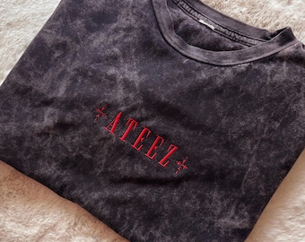 Puede incluir: Una camiseta negra con efecto lavado ácido y cuello redondo. En el centro del pecho presenta la palabra ATEEZ bordada en hilo rojo, flanqueada por pequeños símbolos en forma de cruz roja a ambos lados del texto.