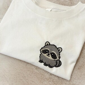 Op de afbeelding: Een cr&egrave;mekleurig T-shirt met korte mouwen en een kleine, geborduurde afbeelding van een schattige, ronde wasbeer aan de rechteronderkant. De wasbeer is afgebeeld in tinten grijs, zwart en wit met grote, donkere ogen en een kleine, lachende mond.