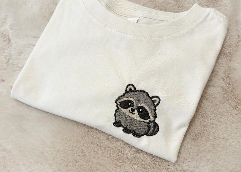 Op de afbeelding: Een cr&egrave;mekleurig T-shirt met korte mouwen en een kleine, geborduurde afbeelding van een schattige, ronde wasbeer aan de rechteronderkant. De wasbeer is afgebeeld in tinten grijs, zwart en wit met grote, donkere ogen en een kleine, lachende mond.