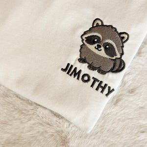 Puede incluir: Una tela de color crema con un dise&ntilde;o bordado de un peque&ntilde;o mapache marr&oacute;n y gris. Debajo del mapache, el nombre JIMOTHY est&aacute; bordado en letras may&uacute;sculas negras y negritas. El bordado est&aacute; detallado con contornos oscuros.