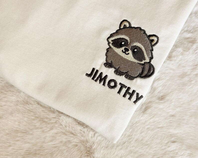 Puede incluir: Una tela de color crema con un dise&ntilde;o bordado de un peque&ntilde;o mapache marr&oacute;n y gris. Debajo del mapache, el nombre JIMOTHY est&aacute; bordado en letras may&uacute;sculas negras y negritas. El bordado est&aacute; detallado con contornos oscuros.