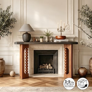 May include: A console table with a dark rectangular top and two light brown wooden legs featuring a vertical row of stacked spheres. The table is positioned in front of a fireplace and holds a gray lamp, a metal candelabra, and small vases.