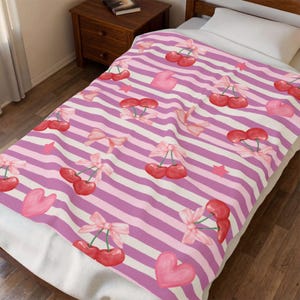 May include: A soft blanket featuring a repeating pattern of red cherries with pink bows, pink hearts, and small pink stars set against a background of horizontal white and light purple stripes. The blanket is draped over a bed with a white pillow.