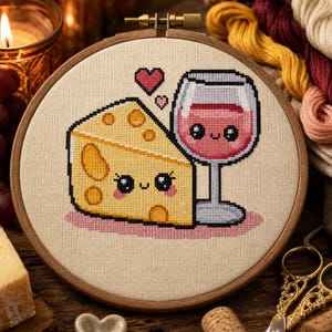 Pode incluir: Um bastidor de bordado circular de madeira contendo um design em ponto cruz sobre tecido bege. O design apresenta uma fatia de queijo amarela com um rosto sorridente e uma taça de vinho rosé com um rosto sorridente, acompanhados por dois corações.