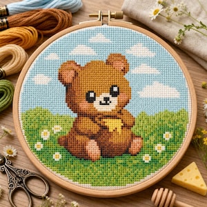 Pode incluir: Um bastidor de bordado circular de madeira contendo um padrão de ponto cruz de um urso castanho sentado num campo de relva verde com flores brancas sob um céu azul claro com nuvens brancas. O urso segura um pote de mel amarelo.