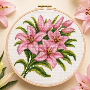 Könnte beinhalten: Ein runder Stickrahmen aus Holz mit weißem Aida-Stoff, der ein Kreuzstichmuster aus rosa Lilienblüten und grünen Blättern zeigt. Das Design umfasst mehrere blühende Lilien sowie geschlossene Knospen, die mittig im Rahmen platziert sind.