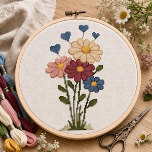 Pode incluir: Um bastidor de bordado circular de madeira contendo um padrão de ponto cruz de um buquê floral com três flores grandes em tons de bege, rosa e vermelho escuro, uma flor azul e vários corações azuis pequenos em um fundo de tecido branco.