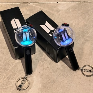 May include: Two black BTS light stick keychains with clear spherical tops containing a glowing blue logo. Each keychain features a black handle with a small logo and a metal ring attachment. They are displayed next to their black rectangular boxes.