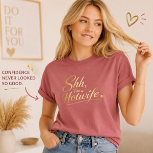 Könnte beinhalten: Ein altrosa T-Shirt mit kurzen Ärmeln und dem goldenen Schreibschrift-Text "Shh, I'm a Hotwife" sowie einem kleinen goldenen Herzsymbol. Das T-Shirt hat einen Rundhalsausschnitt und wird mit einer blauen Jeans kombiniert. Der Aufdruck ist goldfarben.