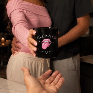 May include: Black coffee mug with the text "CLEAN UP CREW" and a pink tongue graphic. The mug is held by a person wearing a pink off-the-shoulder sweater. The mug is being offered to another person.