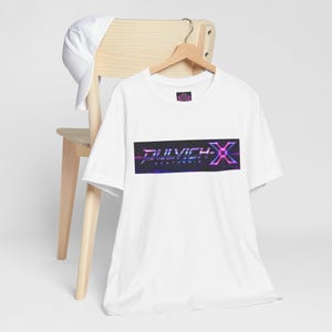 Könnte beinhalten: Weißes T-Shirt mit einem Grafikdesign mit dem Text "DULVICH-X SYNTHESIS" in einem Neon-Stil. Das Design enthält Farbtöne von Lila, Blau und Pink. Eine weiße Baseballkappe liegt auf einem Holzstuhl.