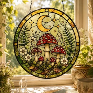 Op de afbeelding: Een ronde glas-in-lood zonnevanger met een centrale groep rood-witte paddenstoelen, omringd door groene varens en kleine witte bloemen. Een gele maansikkel en sterren zijn afgebeeld in het bovenste gedeelte van het glaspaneel.